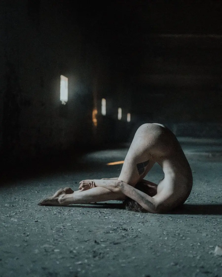 Figure folded into a sculptural pose in a dark industrial space.