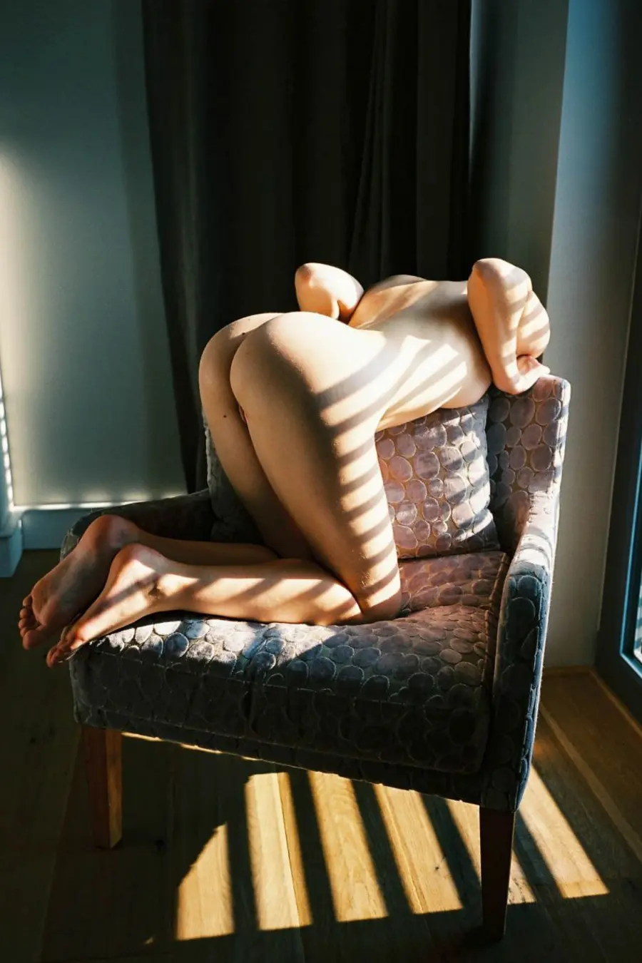 Figure curled into a patterned armchair cut by bands of sunlight.